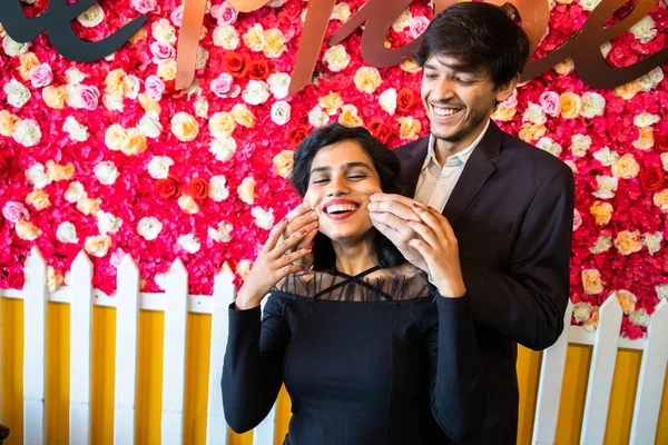 Young happy cheerful indian couple in love have fun together, man pinching cheeks of his girlfriend against floral background, enjoy life, new year or valentines day.