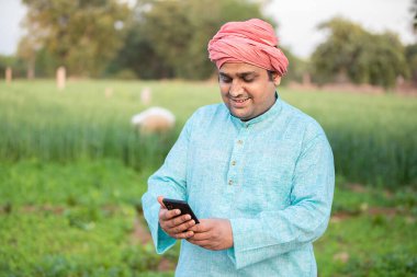 Genç mutlu Hintli çiftçi akıllı telefon kullanarak tarım yeşil tarlasında, internet bankacılığı, 5G ağ teknolojisi konsepti, geleneksel kurta kıyafeti giyen erkek.