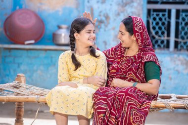 Gülümseyen genç Hintli anne ve sevimli küçük kız birlikte vakit geçirmekten zevk alıyor örgülü saçlı tatlı kız ve kırmızı sarili anne birbirine bakıyor, geleneksel yatakta oturuyor..