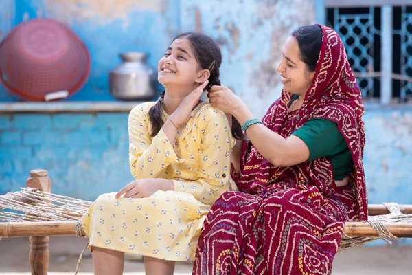 Rural indian mother braids hair of her young adorable daughter, cute girl feel pain as mum getting her ready sitting on traditional bed at village home. Both spending time together.