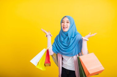 Asyalı Müslüman Arap, mutlu genç kadın portresi İslam dini peçeli komik bir tebessüm takıyor elinde renkli alışveriş torbaları tutuyor mutlu alışveriş torbaları sarı stüdyoda el kaldırıyor