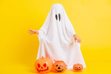 Komik Cadılar Bayramı Çocuğu Kavramı, beyaz kostümlü, cadılar bayramı hayaleti gibi tatlı bir çocuğun tüm vücudu elinde turuncu kabak hayaleti, beyaz arka planda sarı bir stüdyo fotoğrafı.