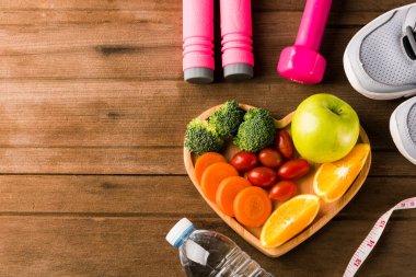 Kalp tabağı odunundaki taze meyve ve sebzelerin üst görüntüsü (elma, havuç, domates, portakal, brokoli) ve dambıl, ahşap masadaki spor ayakkabıları, sağlıklı yaşam tarzı diyet gıda konsepti