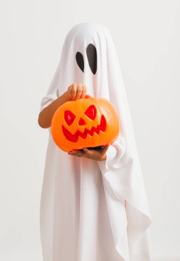 Komik Cadılar Bayramı Çocuğu kavramı, beyaz kostümlü küçük şirin çocuk Cadılar Bayramı hayaleti korkunç elinde turuncu kabak hayaleti, beyaz arka planda tek başına çekilmiş bir stüdyo resmi.