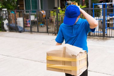 Mavi üniformalı Asyalı genç teslimatçı. Hasarlı karton kutuyu elinde tutuyor. Evin önünde kırılıyor.