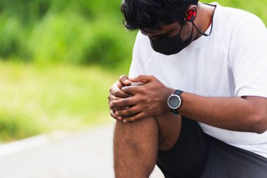 Kapatın Asyalı genç spor koşucusu. Siyah adam, el ele oturuyor. Açık hava sokak sağlık parkında koşarken diz ağrısı çekiyor.