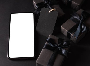 Kara Cuma alışverişi konsepti, hediye kutusunun üst görüntüsü siyah kağıt ve siyah kurdele sarılı akıllı telefon boş ekran, karanlık arka planda stüdyo çekimi