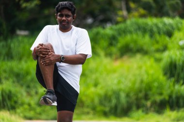 Asyalı genç atlet spor koşucusu ayakta duran siyahi adam koşmadan önce ayak ısıtması giyer. Açık havada koşmak için tek dizini göğsüne kadar gerer ve sağlıklı egzersiz yapar.