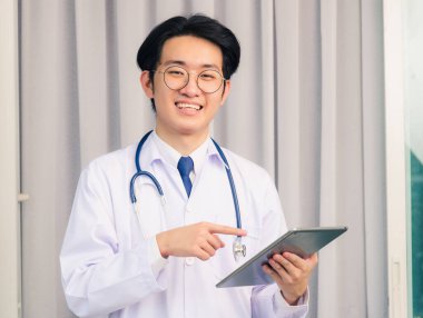 Mutlu Asyalı genç doktorun portresi üniformalı, steteskopla gülümseyen yakışıklı adamın boynu bağlanmış, akıllı dijital tableti gösteren ve kameraya bakan, sağlık sigortası konsepti