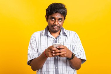 Asyalı mutlu bir portre genç siyah adam ayakta gülümsüyor cep telefonuyla sosyal mesaj yazarken veya okurken cep telefonu boş ekranında, stüdyo sarı arka planda fotokopi alanı ile çekim yapıyor.