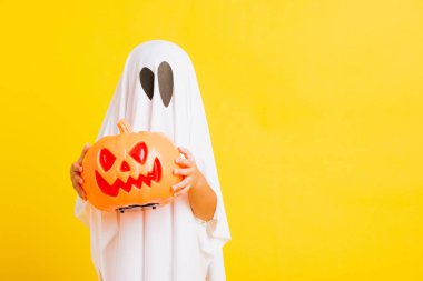 Komik cadılar bayramı çocuğu kavramı, beyaz kostümlü şirin çocuk Cadılar Bayramı hayaleti korkunç elinde turuncu kabak hayaleti, beyaz arka planda sarı bir stüdyo.