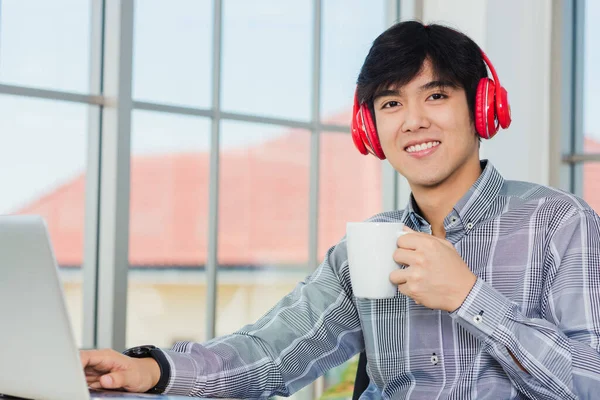 アジアの若いビジネスマンの幸福と赤いヘッドフォンノートパソコンとデスクの職場の自宅のオフィスに座って 自信を持ってハンサムな男のライフスタイルの笑顔は コーヒーカップを保持リラックス音楽に耳を傾ける — ストック写真