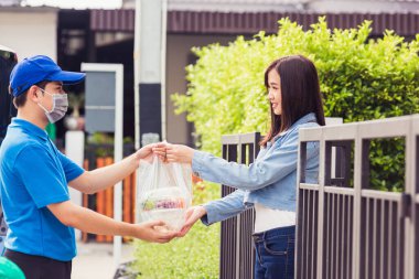 Asyalı genç teslimatçı yüz maskesi takıyor. Bakkal servisi yapıyor. Kadın müşterilere plastik torbalar veriyor.