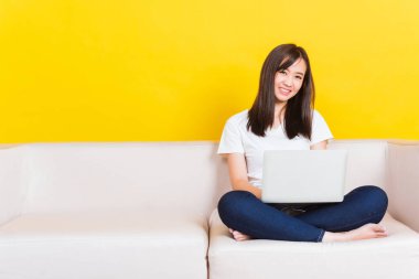 Asyalı mutlu bir genç kadının portresi. Kanepede oturuyor. Oturma odasındaki dizüstü bilgisayarla sarı arka planda izole edilmiş.