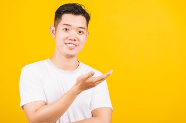 Portre mutlu Asyalı yakışıklı adam beyaz tişört giyerek ayakta duruyor elinde kameraya bakan bir şey var, stüdyo görüntüsü izole edilmiş sarı arka plan.