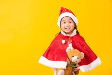 Noel Baba şapkalı çocuk, gülümseyen Asyalı küçük tatlı kızın portresi, Noel 'de oyuncak ayıya sarılma konsepti, stüdyo resmi, sarı arka planda izole edilmiş, mesaj alanı için fotokopi alanı.