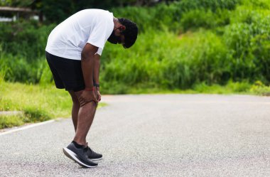 Asyalı genç sporcu siyah adam ayakta duruyor. Ayaklarını giyip açık havada koşuyor. Koşarken dizini tutuyor.