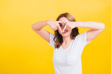 Asya Tayland mutlu portresi güzel güzel genç kadın gülümsemesi parmak kalp figürü şekil işareti yap iki elle ve kamerayla işaret et, stüdyo görüntüsü izole edilmiş sarı arka plan fotokopi alanı ile