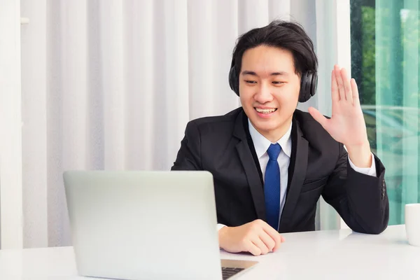 Work from home, Asian young businessman smile wearing headphones and suit video conference call or facetime on desk and raise your hand to say hello greet colleague at home office