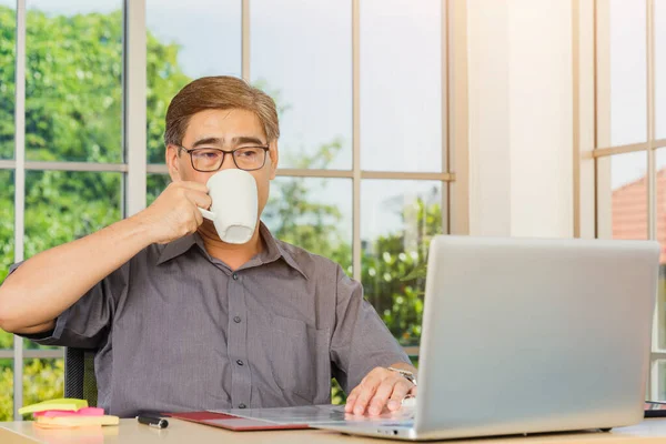 事務所のあるデスクオフィスに座っているアジアの上級ビジネスマン 職場の自宅のオフィスでラップトップコンピュータを使用して自信を持っている中年のハンサムな男 そしてコーヒーを飲みます — ストック写真