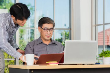 Asyalı kıdemli ve kıdemsiz iki işadamı, danışma projeleri sırasında iş ortağıyla birlikte masa başı bilgisayarında çalışan olgun bir patron hakkında tartışıyorlar.
