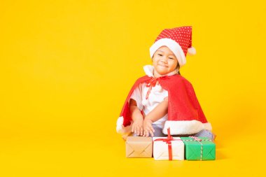 Asyalı küçük şirin kız gülümseyen surat, kırmızı Noel Baba şapkalı bir çocuk Noel tatili konsepti ya da yeni yılın mutlu noelleri, sarı arka planda izole bir şekilde oturuyor.