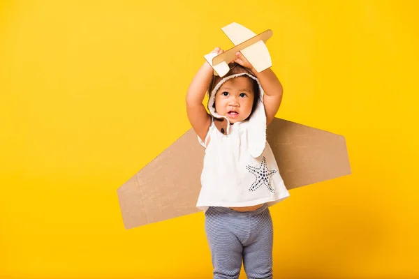 幸せなアジアの美しい面白い赤ちゃんの女の子の笑顔は おもちゃの段ボール飛行機の翼を持つパイロットの帽子の遊びやゴーグルを身に着けている飛行機のおもちゃを保持飛ぶ スタジオショット孤立黄色の背景 スタートアップの自由コンセプト — ストック写真