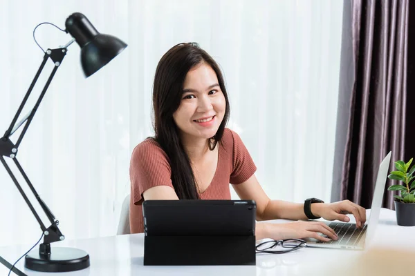 Work from home, Smiling Happy Asian business young beautiful woman sitting on desk workspace use modern smart tablet and laptop computer technology video call conferencing to team at home office