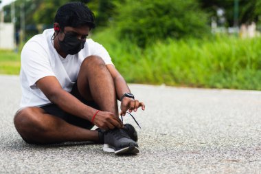 Asyalı spor koşucusu yüz maskesi takıyor. Ayakkabı bağcığı takıyor. Koşu ayakkabılarını giymeye çalışıyor.