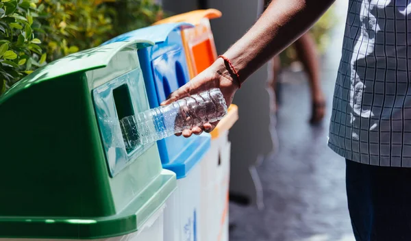 Selective focus close up the man black hand throwing an empty plastic water bottle in the recycling garbage trash or bin, environmental recycling concept