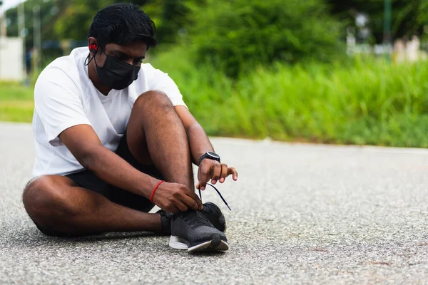Asyalı spor koşucusu yüz maskesi takıyor. Ayakkabı bağcığı takıyor. Koşu ayakkabılarını giymeye çalışıyor.