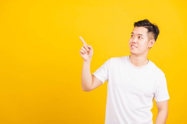 Mutlu Asyalı yakışıklı adam gülümsüyor beyaz t-shirt giyip uzayı işaret ediyor. Stüdyo resmi, izole edilmiş sarı arka plan.