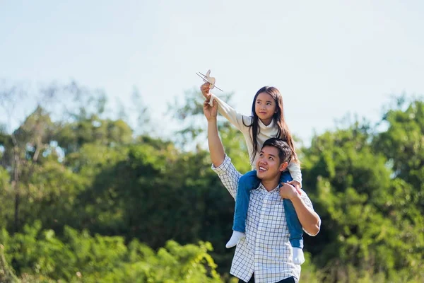 Happy Asian young family father and carrying an excited girl on shoulders having fun and enjoying outdoor lifestyle together playing aircraft toy on sunny summer day, Father\'s day concept