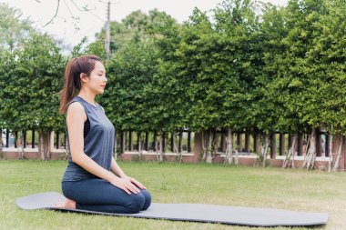 Meditasyon pozisyonunda yoga yapan Asyalı genç bir kadın yeşil çimlerin üzerinde otururken gözleri kapalı bir bahçe parkı, esneme, meditasyon, egzersiz sağlık konsepti.