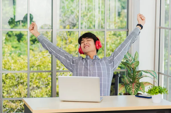 アジアの若いビジネスマンは 自宅のオフィスで赤いヘッドフォンで音楽を聞いて笑顔 幸せなビジネスマンは 机の上に座って リラックスした着用のワイヤレスヘッドフォンは 音楽を聴くとラップトップコンピュータで作業を楽しむ — ストック写真