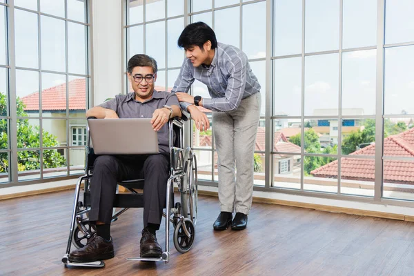 Asian senior disabled businessman in a wheelchair with laptop computer discuss together with team in office. Old father man sitting wheelchair and his son talking video calls conference on laptop