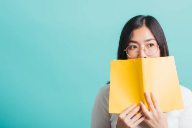 Genç ve güzel Asyalı bir kadın açık bir kitabın arkasına saklanıyor. Gözlüklü bir kadın, elinde kitap tutuyor ve okuyor. Stüdyo resmi, mavi arka planda, eğitim konseptinde izole edilmiş.