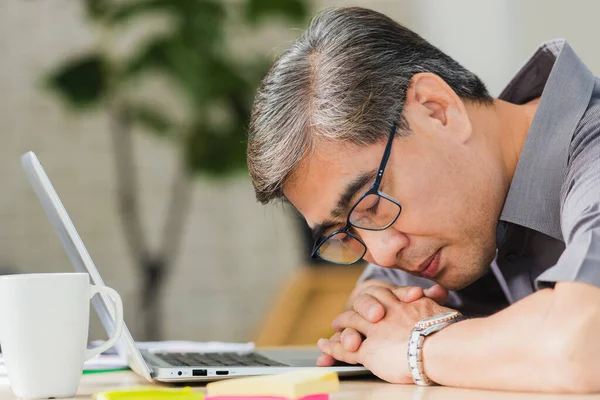 アジアのビジネスマンは彼が机の上のノートパソコンの上で寝て過労した 職場のテーブルの上に寝そべって眼鏡をかけた先輩が — ストック写真