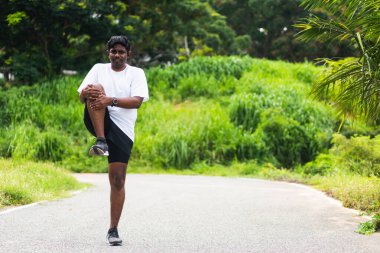 Asyalı genç atlet spor koşucusu ayakta duran siyahi adam koşmadan önce ayak ısıtması giyer. Açık havada koşmak için tek dizini göğsüne kadar gerer ve sağlıklı egzersiz yapar.