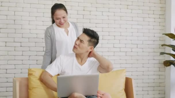 幸せなアジアの家族のカップルの夫と妻は自宅から働くラップトップコンピュータを使用してソファに座って笑っている 男座っています上のソファホーム作業上のラップトップ彼女の女性あります与えます彼女の肩マッサージ — ストック動画
