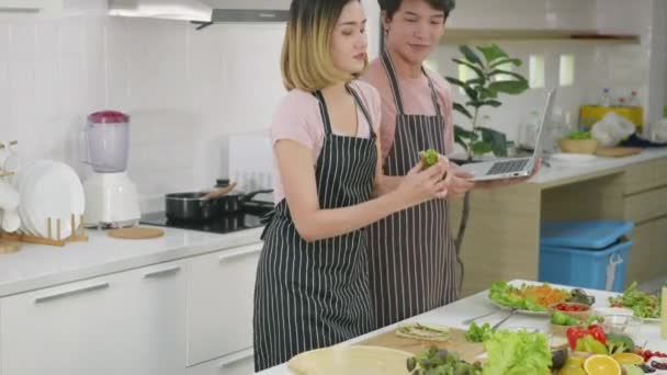 幸せなアジアの美しい若い家族のカップルの夫と妻は台所で一緒に家庭で野菜サラダを調理します 男性と女性は ラップトップコンピュータ上のレシピにオンライン調理クラスによると — ストック動画