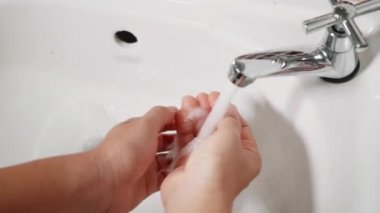 Genç kadın ellerini sabun ve musluğun altında yıkayarak kapatıyor. Kadın avuçlarını otel banyosundaki lavaboda beyaz köpüklerle yıkıyor. Hijyen onu Covid-19 'dan koruyor.