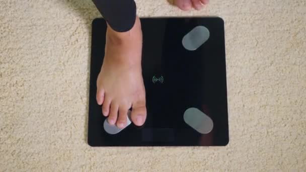 アジアの若い女性の足の床の電気スケールに立って 女性は自宅でリビングルームで働いて 女性はデジタル計量機の上に立つ 健康的な減量コントロールの概念 — ストック動画