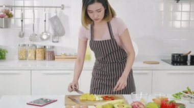Mutfaktaki mutlu Asyalı güzel kadın dijital tablet bilgisayarda tarifi takip ediyor. İnternet sitesindeki internet teknolojisindeki tablet içeriği üzerinden kadın pişirme salatası çevrimiçi video klibi. modern yiyecek