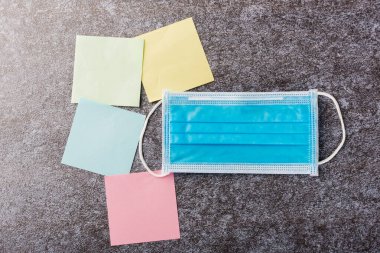 Boş kağıt çubuk listesinin üzerinde sarı, yeşil ve mavi renkli yüz maskesi olan beton arka planda, karantina hastalığı koronavirüsü ya da COVID-19 ve not çubuğu belleği çalışması bugün