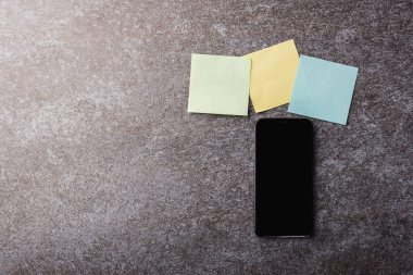 Boş kağıt yapıştırma listesinin üzerinde sarı, yeşil ve mavi renkli modern akıllı cep telefonu boş ekranlı beton arka plan, iş hatırlatma konsepti