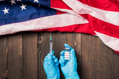 Ellerinde Coronavirus (COVID-19) aşı şişesi ve Amerika Birleşik Devletleri bayraklı şırınga bulunan ahşap kaplama eldivenleri olan bir doktorun elleri, ABD Aşısı