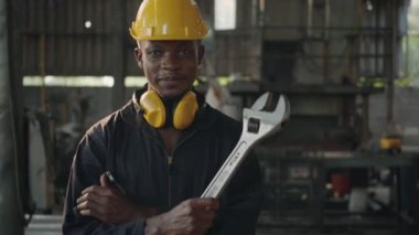 Ön makinede sarı başlıklı gülümseyen Amerikalı endüstriyel işçi, İngiliz anahtarı ve kollarını fabrikada çapraz tutan mühendis..