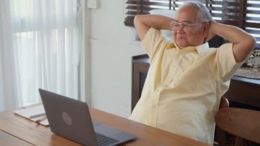Üst düzey adam gözlük takıyor, sandalyede oturuyor evdeki dizüstü bilgisayarda çalışıyor, mutlu yaşlı adam bilgisayarla emekli oluyor, yaşlı büyükbaba evde çalışıyor, ağır çekimde.