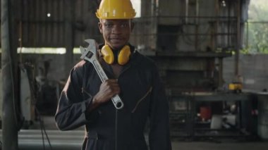 Ön makinede miğfer ve kulak koruyucusuyla gülümseyen Amerikalı endüstriyel işçi, fabrikada çalışırken omzunda ingiliz anahtarı tutan mühendis..